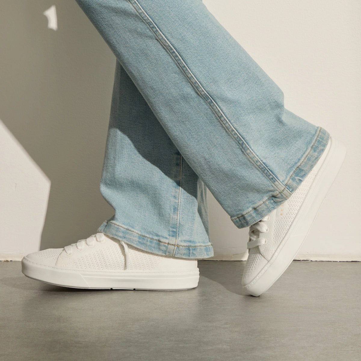 White knit sneakers facing left, worn with light blue jeans on a concrete floor