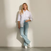 Smiling woman in a white shirt and jeans wearing white sneakers, facing forward against a beige wall