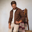 Man seated wearing a brown Coltrane Pique Shirt, beige pants, next to a patterned woven blanket on a wooden chair