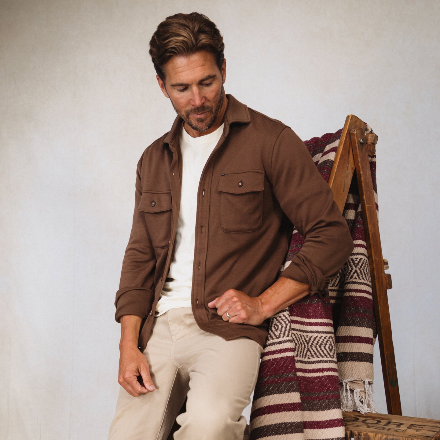Man seated wearing a brown Coltrane Pique Shirt, beige pants, next to a patterned woven blanket on a wooden chair