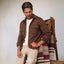 Man seated wearing a brown Coltrane Pique Shirt, beige pants, next to a patterned woven blanket on a wooden chair