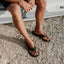 Fanning flip flops facing forward on feet, showing black synthetic upper and cushioned footbed with arch support by the beach.