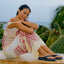 Woman sitting outdoors wearing Sunset Low Maya black sandals facing right, showing side and top views with a tropical background