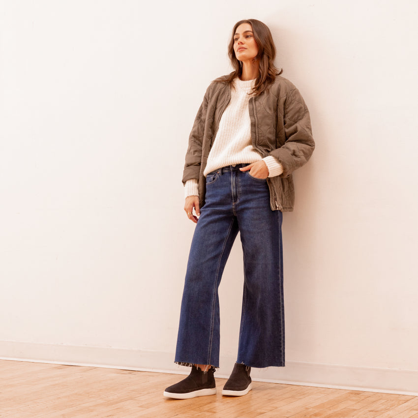 Woman standing wearing Neptune Athena black slip-on shoes facing left, styled with casual denim and jacket.
