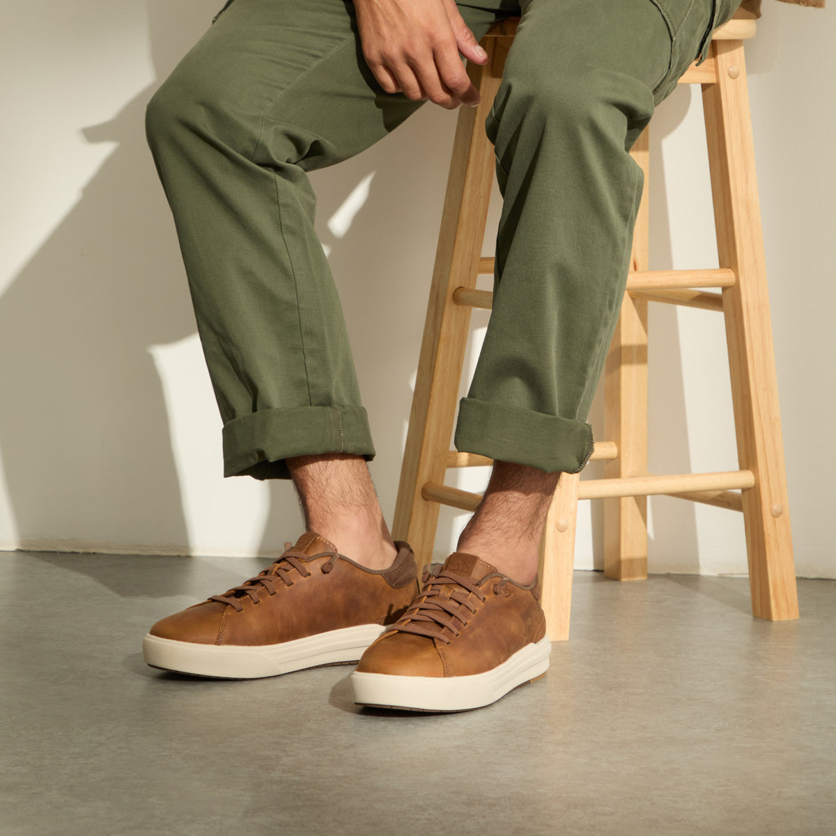 Brown lace-up sneakers facing left, worn with rolled olive pants while seated on a wooden stool