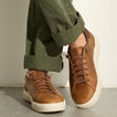 Brown leather sneakers facing forward, paired with olive green pants on a concrete floor