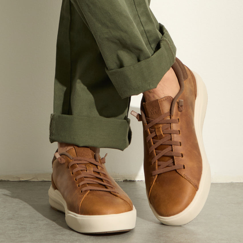Brown leather sneakers facing forward, paired with olive green pants on a concrete floor