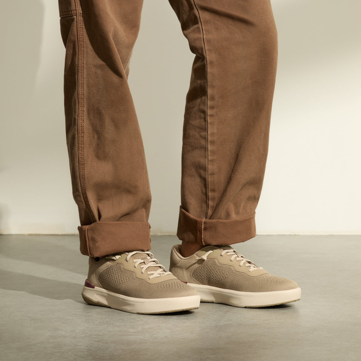 Beige lace-up sneakers facing right, worn with cuffed brown pants on a concrete floor