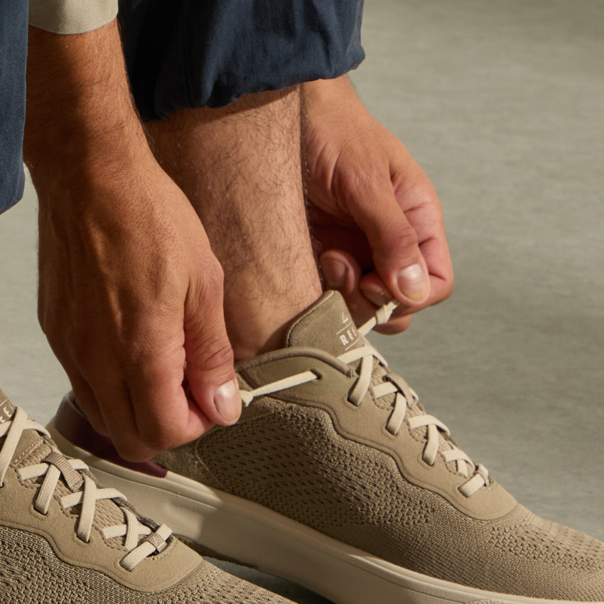 Close up of hands tying the laces on a beige sneaker facing right with a textured upper and cushioned sole
