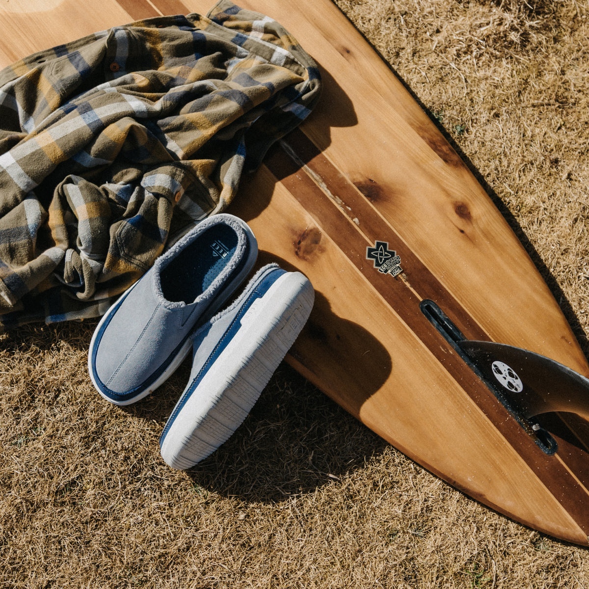 Gray slip-on shoes facing lower left beside a wooden surfboard and plaid shirt on dry grass