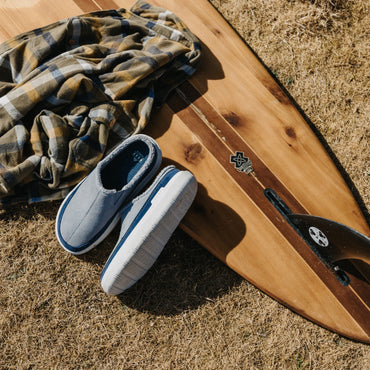 Gray slip-on shoes facing lower left beside a wooden surfboard and plaid shirt on dry grass
