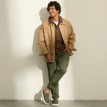 Man in casual apparel wearing olive sneakers with toes facing forward, posed against a wall.