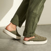 Olive green slip-on shoes facing right, worn with cuffed pants on a concrete floor
