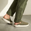 Olive green slip-on shoes facing right, worn with cuffed pants on a concrete floor