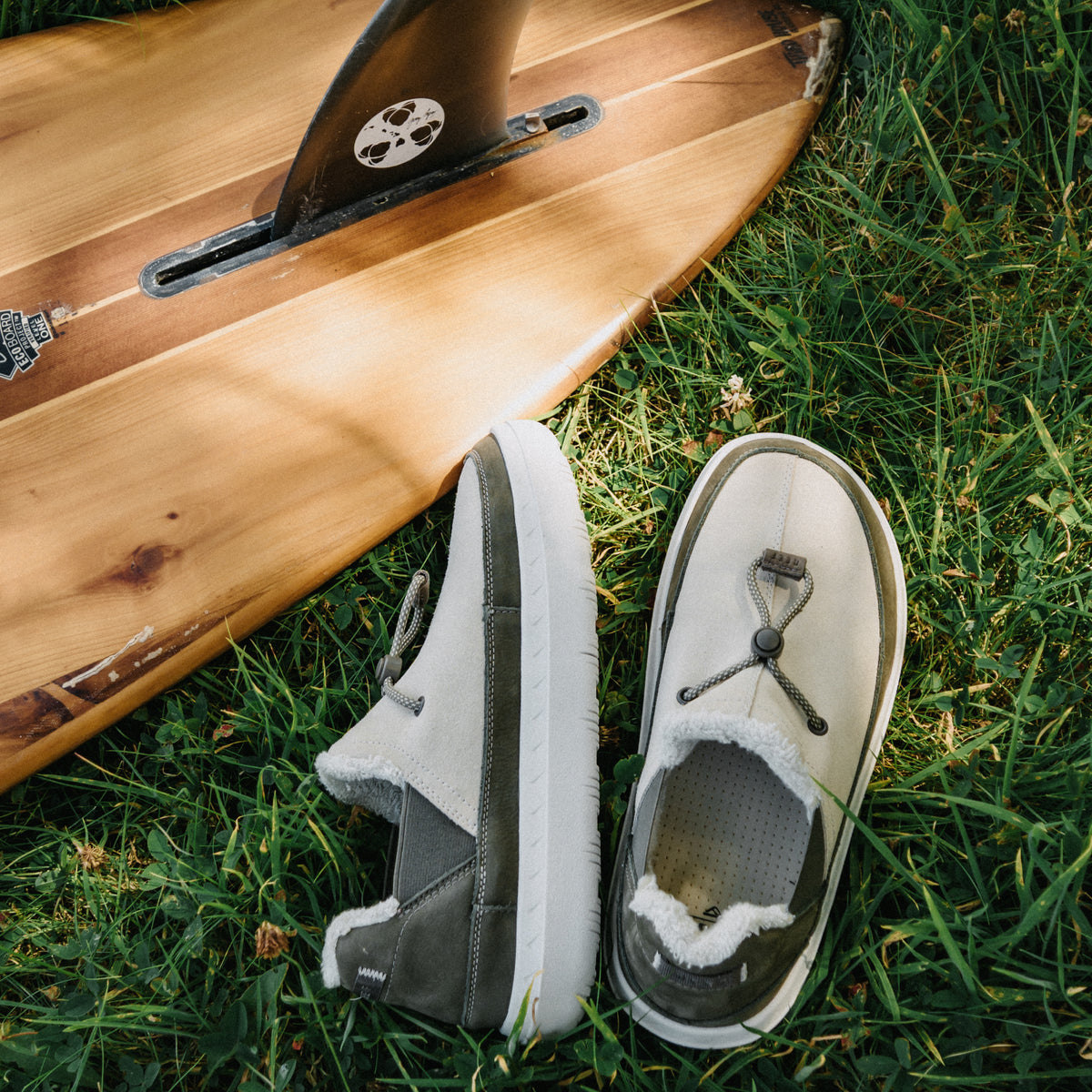 REEF Rambler shoes facing upper left and upper right beside a surfboard on grass with cozy lining and cushioned soles