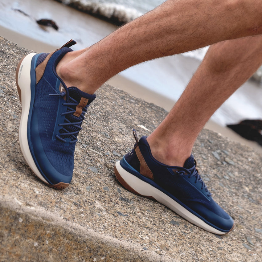 Reef Zuma navy sneakers, one facing left and one facing right, worn on a beachside walkway for everyday comfort