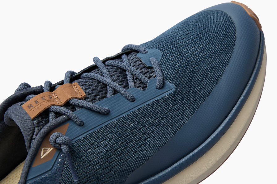 Close-up of a navy lace-up shoe facing upper right, showing a textured upper, cushioned sole, and brown accents