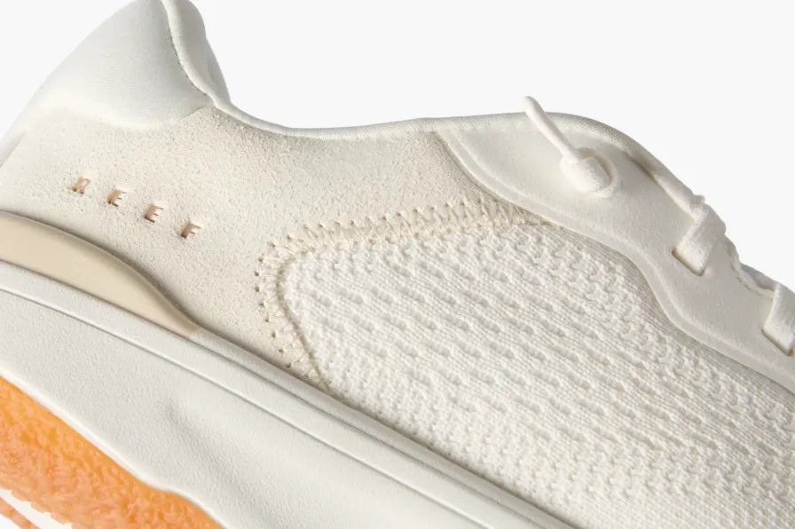Close-up of the left side of a white REEF sneaker highlighting textured knit upper and cushioned sole with orange outsole edge.