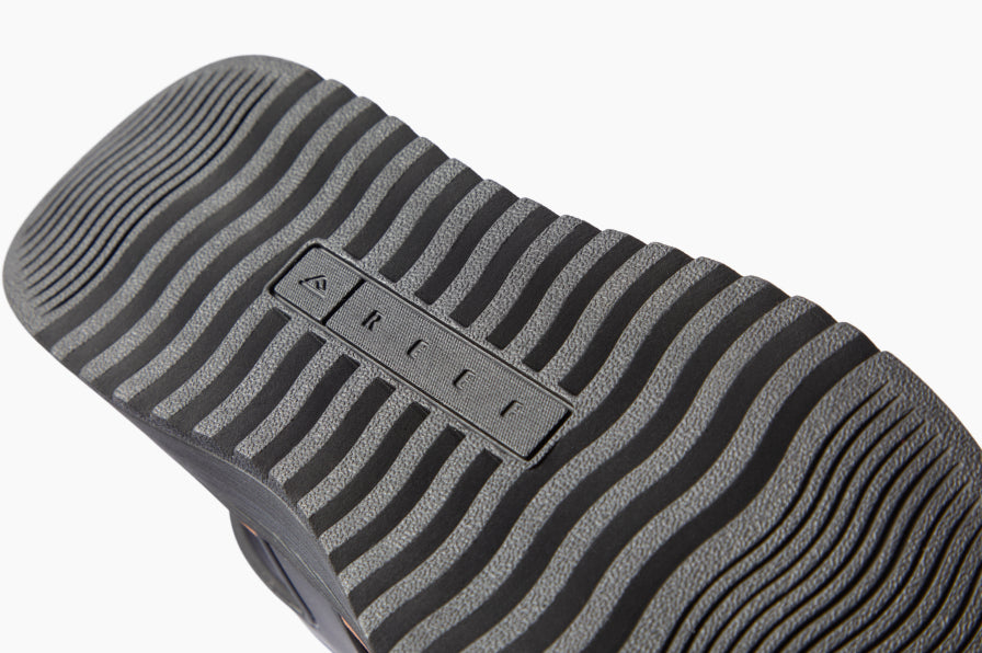 Close-up of a black and gray REEF shoe outsole facing diagonally from lower left to upper right, showing wavy traction grooves.