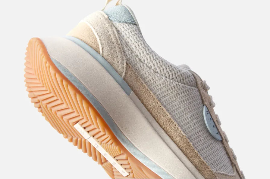 Side view of beige and light blue sneaker facing right, showing textured knit upper, cushioned midsole, and patterned outsole.