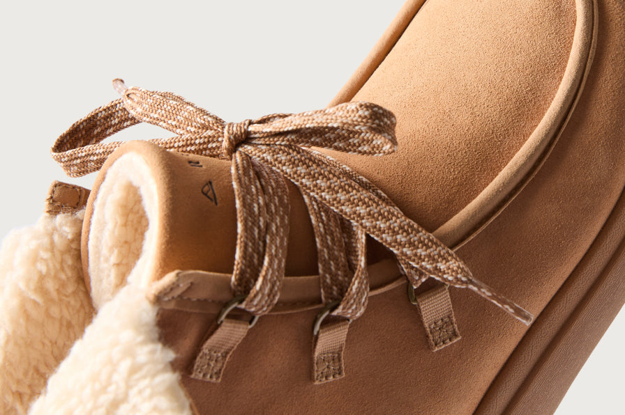 Close-up of tan suede boot facing left, showing tied laces and plush cream-colored shearling collar for coastal comfort.