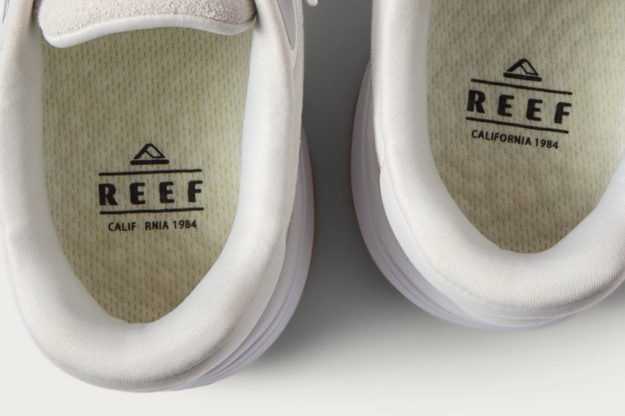 Close-up of white REEF shoes facing upward, showing cushioned insoles with branded footbeds