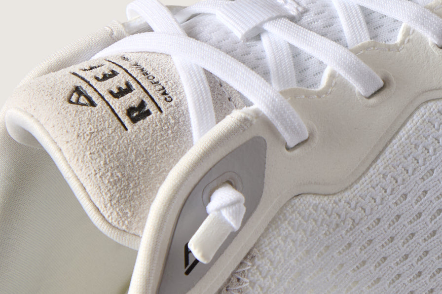 Close-up of a white REEF sneaker facing left, showing its logoed tongue, laces, and textured knit upper.