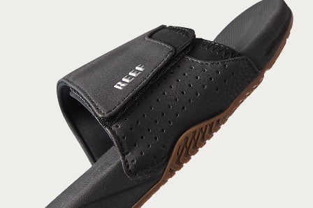 Close-up of a black REEF slide sandal facing right with adjustable strap and textured brown sole visible beneath.