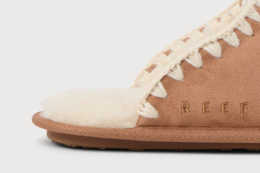 Close-up of the left side of a tan slip-on shoe with suede upper and plush lining showcasing stitched trim and REEF branding.