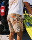 Man wearing Men's Palmera Boardshort - Floral Camo standing by truck on beach holding yellow surfboard in sunlight