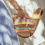 Woman holding a pair of Cushion Vista sandals facing up, featuring multicolor straps and cork footbeds for beach comfort.