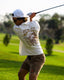 Man swinging golf club wearing High-Stoke Relaxed Tee - Bone with brown shorts, casual beach-inspired athletic look outdoors