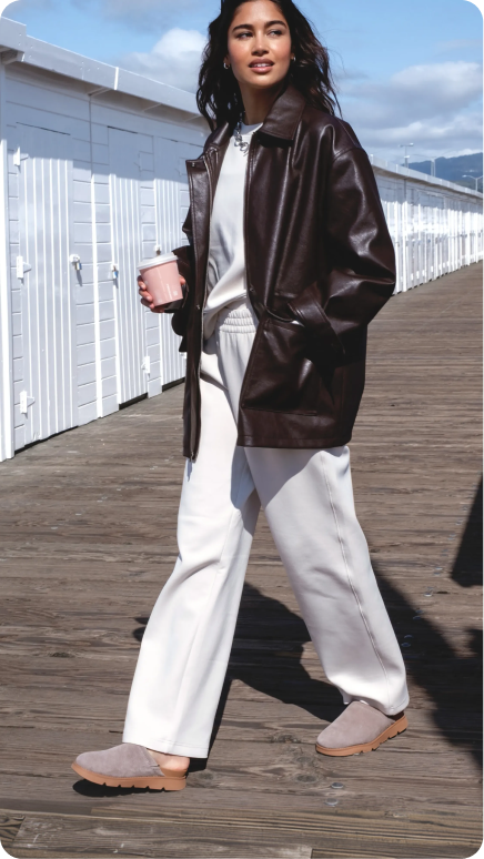 Side view of a person walking on a boardwalk wearing light brown clogs with cushioned footbed and white pants, facing right.