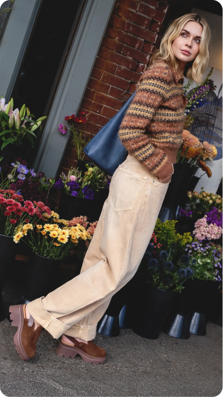 Side view of woman wearing brown leather clogs with chunky sole and cushioned footbed, standing by flower shop.