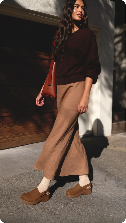Side view of person walking outdoors wearing brown slip-on clogs with cushioned sole, cream socks, and casual beige pants