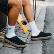Person wearing black slip-on shoes, one facing left and the other right, beside a skateboard on stone pavement