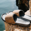 Tailslide tan men's slides facing right with a close up view of padded straps and contoured footbeds on a rocky beach