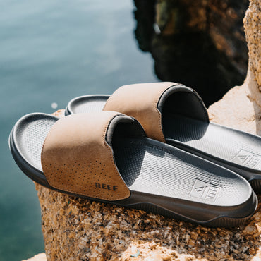 Tailslide tan men's slides facing right with a close up view of padded straps and contoured footbeds on a rocky beach