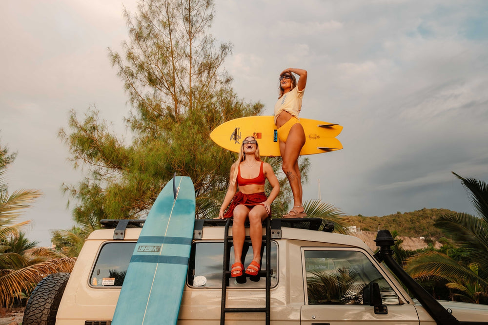 Two women pose atop a camper van with surfboards while the seated woman wears red sandals facing forward