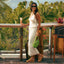 Woman wearing Vista Higher Cleo tan suede sandals facing left, standing outdoors with tropical foliage background.