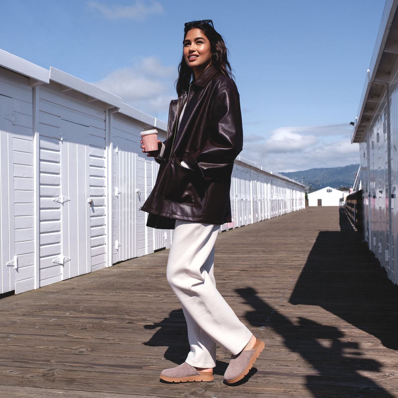 Woman standing on boardwalk facing right wearing Vista Luxe Mariana brown suede clogs with arch support and casual white pants.