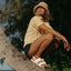Woman wearing Reef Water Vista white and tan sandals facing right, sitting on a tree trunk at the beach, showing front and side view.