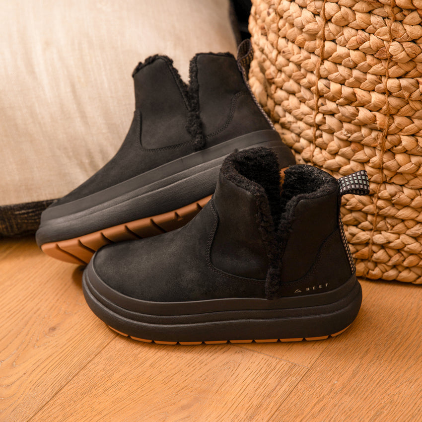 Two black ankle boots with fleece lining and textured soles facing left on a wood floor