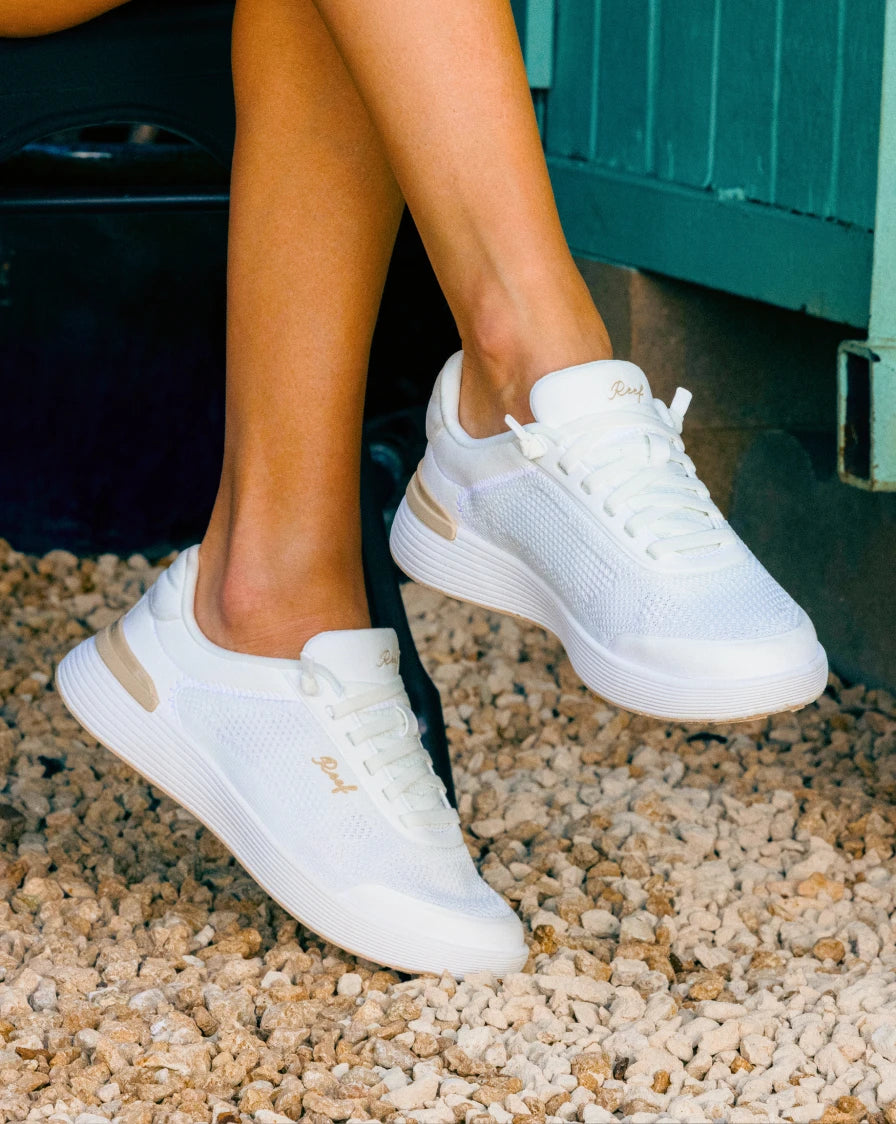 Lower legs of a person sitting with her legs crossed and wearing white lace-up sneakers with some small rocks on the floor
