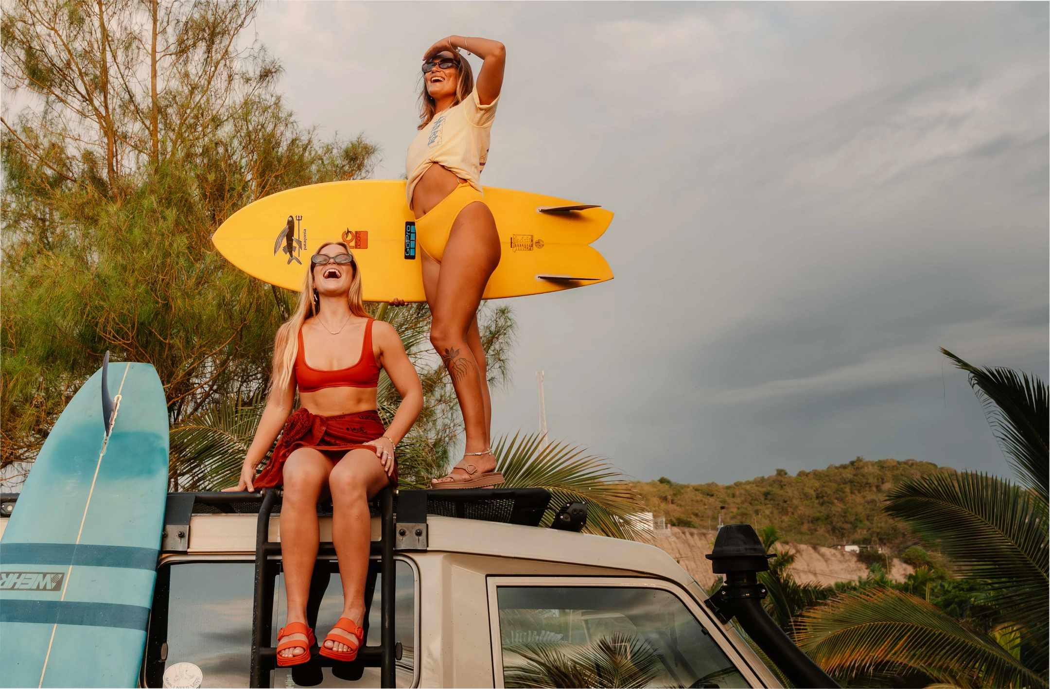 Two women on top of a car outside with surfboards, smiling and wearing red and tan colored two-bar slide sandals