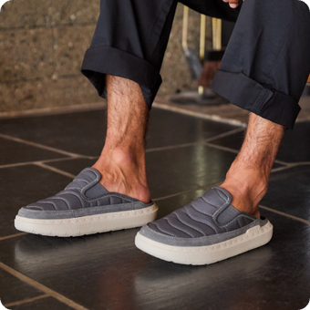 Gray slip on shoes facing left on a persons feet showing cushioned soles and stitched uppers