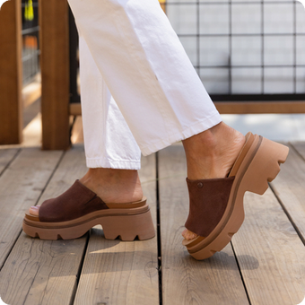 Close up of brown clogs facing left, showing side profiles and chunky soles on a wooden deck