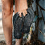 Hand holding black Seaside Twist sandals facing down with water splashing, showcasing comfort and beach-ready design.