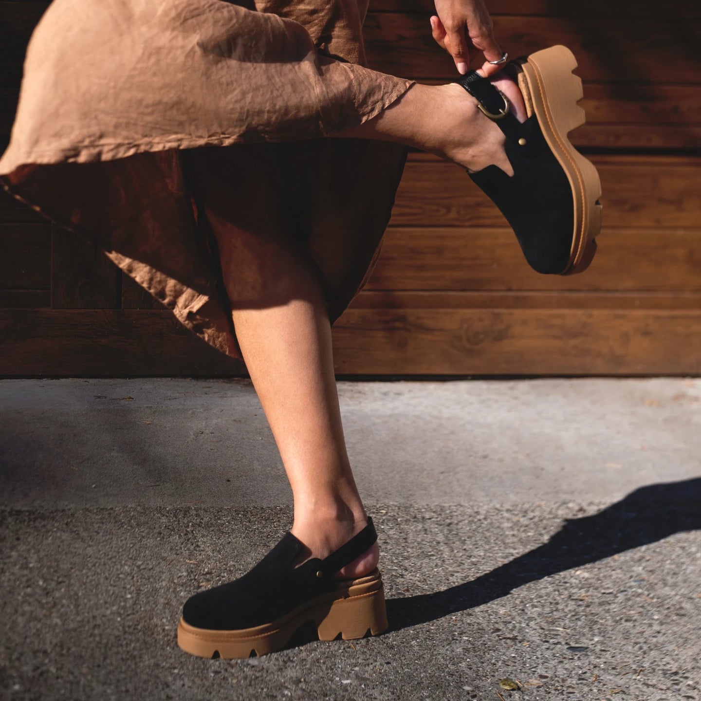 Woman adjusting left-foot black suede Vista Luxe Solene clog showing side and sole, platform heel in outdoor setting