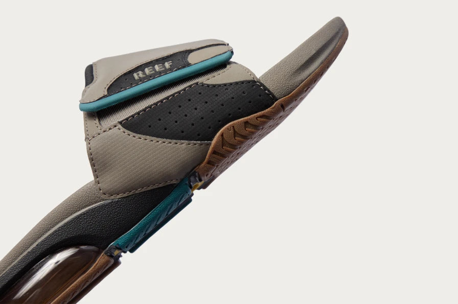 Side view of grey and black slide sandal angled upward, showing cushioned footbed and textured sole edge with REEF logo.